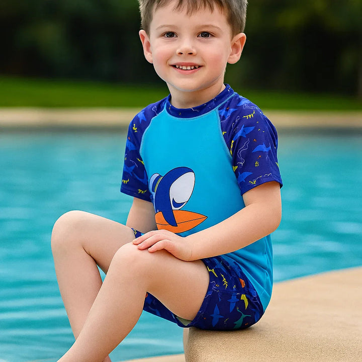 Child sitting beside a pool in a short-sleeve shark swim shirt and matching shorts
