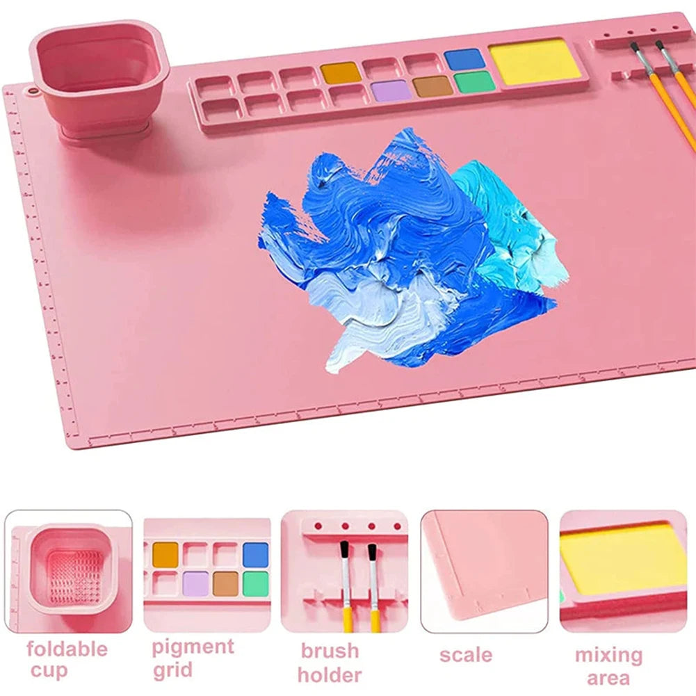Pink painting mat showing a folding cup, paint wells, brush holders, edge ruler, and mixing area.