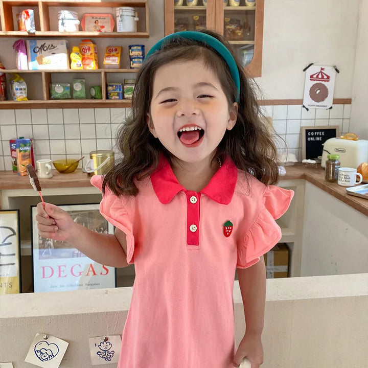Front view of a child wearing the pink dress with a contrasting collar and strawberry detail