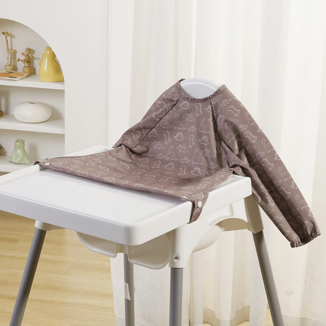 Taupe long-sleeve baby bib with outlined dinosaurs on a high chair