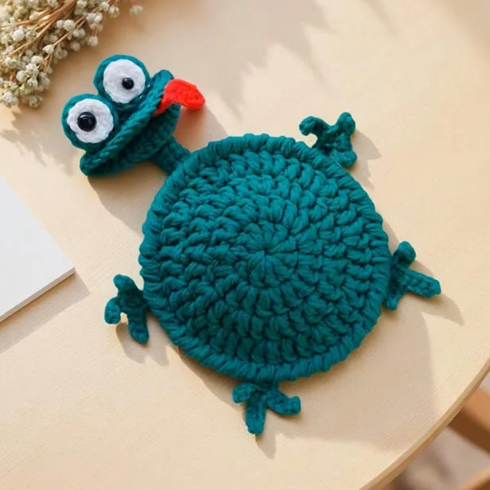 Teal crocheted frog coaster with large eyes, little feet, and a red tongue