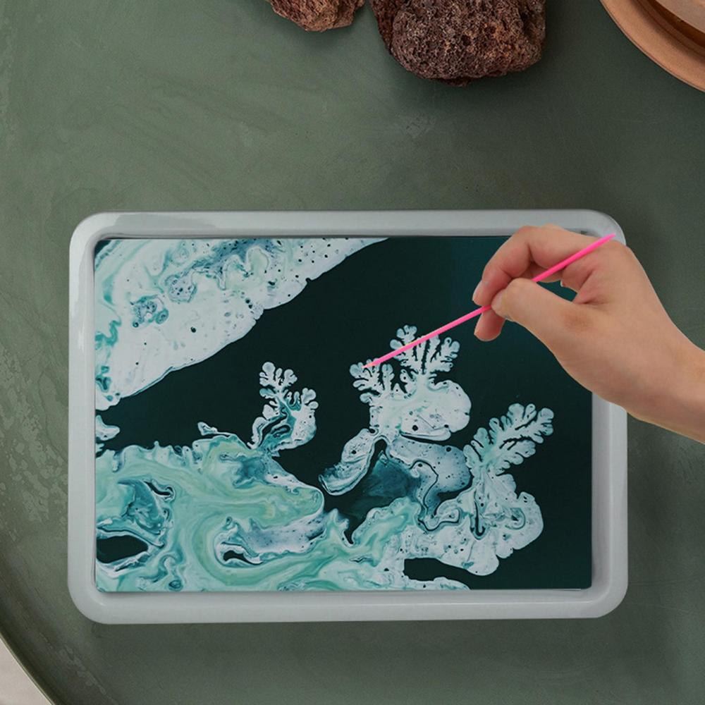 A hand shaping teal and white marbling patterns in a shallow tray with a pink stick