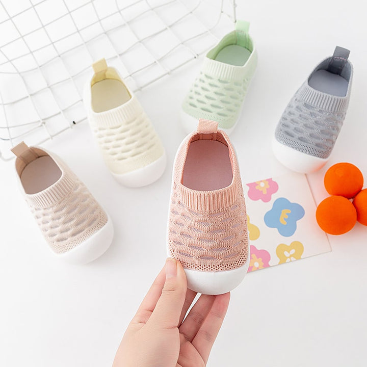 Hand holding a pink knit shoe with beige, yellow, green, and gray shoes behind it