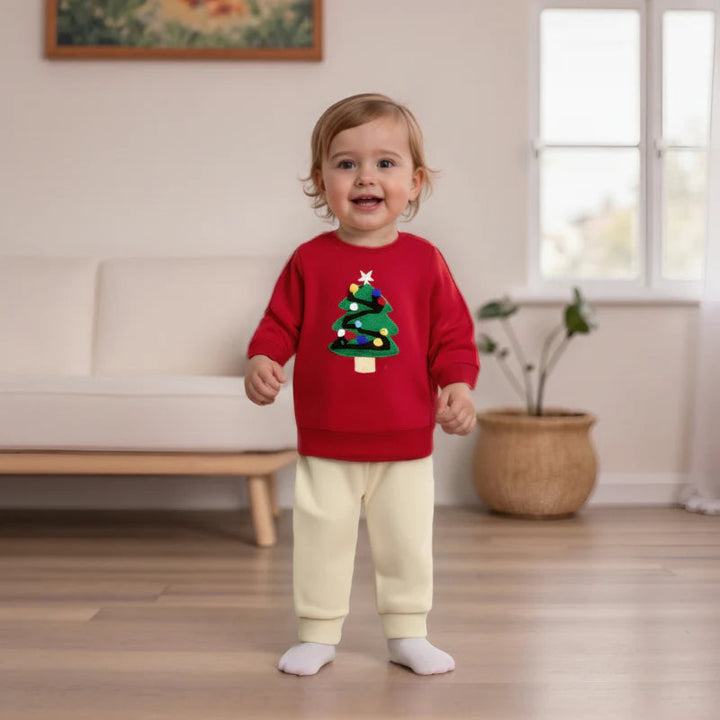 Toddler wearing a red Christmas tree sweatshirt and cream pants in a living room.