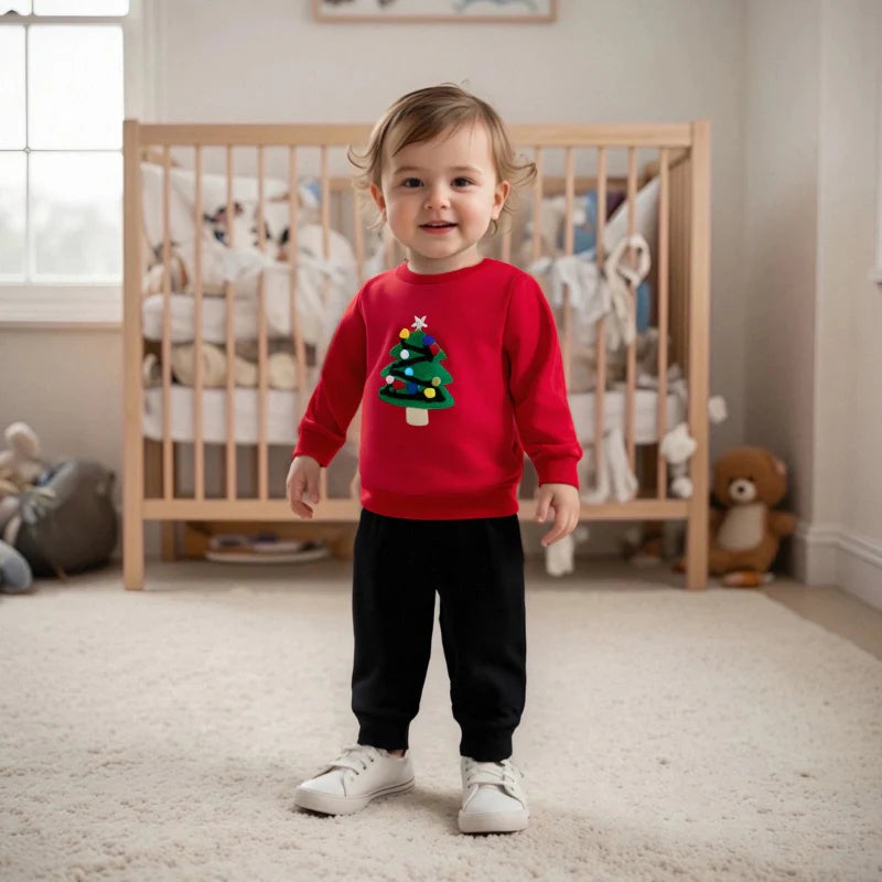 Toddler in a red Christmas tree sweatshirt, black pants, and white sneakers in front of a wooden crib.