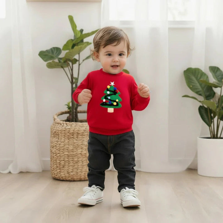 Toddler wearing a red Christmas tree sweatshirt with dark pants and white sneakers beside indoor plants.