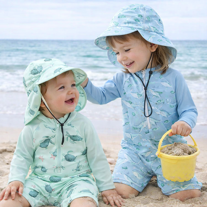 Two children in mint green turtle and light blue whale swim sets with matching hats at the beach.