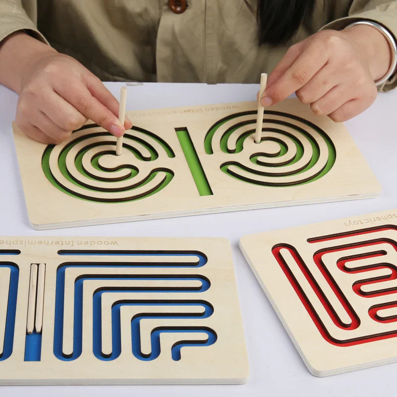 Two hands guide styluses through a green spiral maze, with blue and red boards nearby