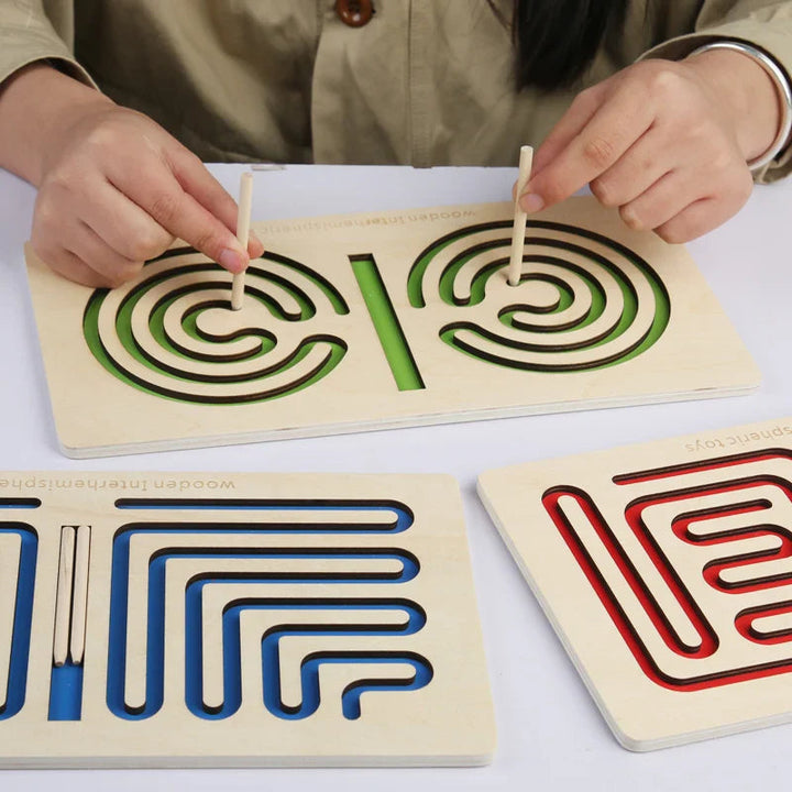 Two hands guide styluses through a green spiral maze, with blue and red boards nearby