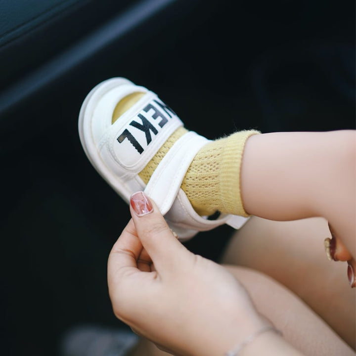 An adult adjusts the ankle strap of a white baby sandal worn over a yellow sock