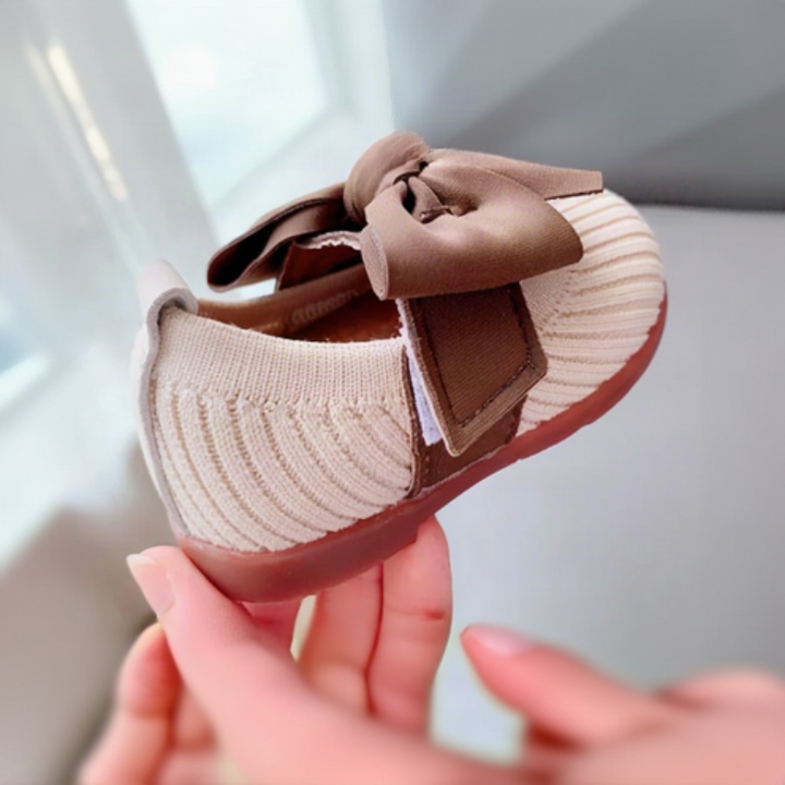 Side view of a white knitted baby shoe with a brown strap and pink rubber sole