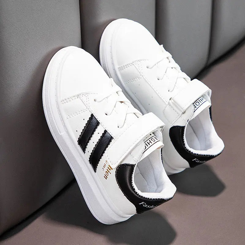 Pair of white kids' sneakers with black accents and top straps resting against a gray sofa.