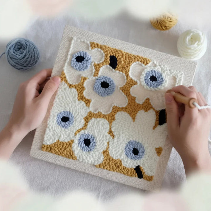 Hands stitching white flowers with blue centers on a mustard-colored embroidery background.