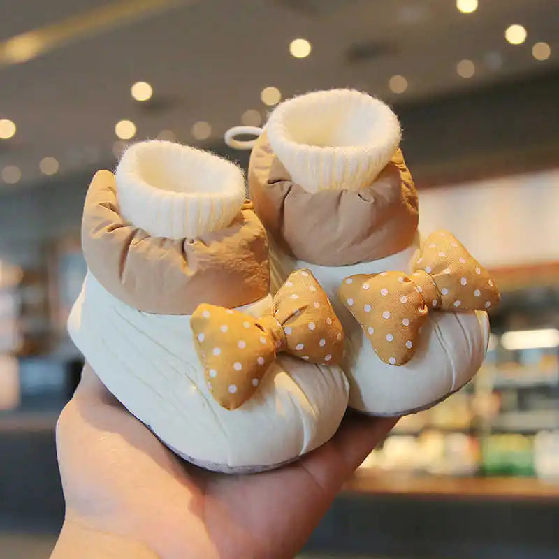 White baby shoes with tan upper panels, cream ribbed cuffs, and dotted bows, held in one hand