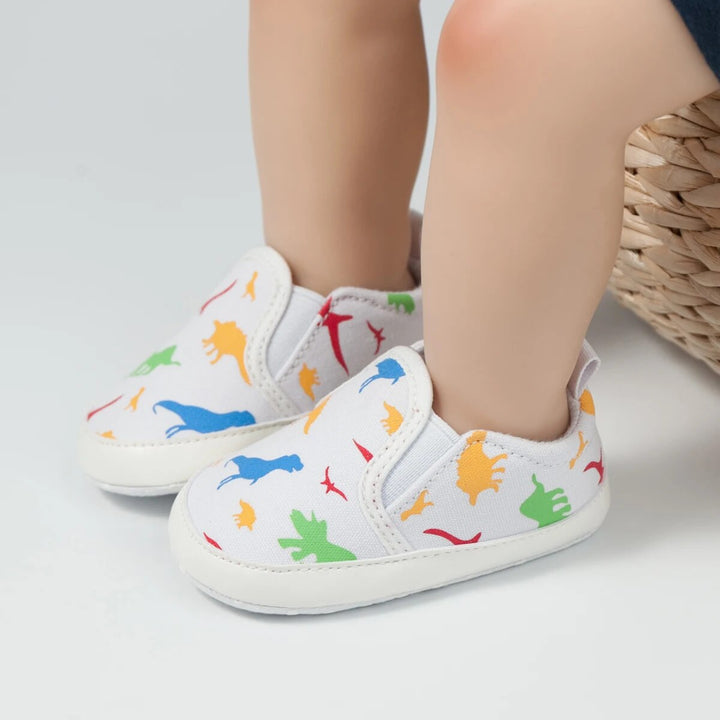 White baby slip-on sneakers with multicolor dinosaur prints, shown on feet