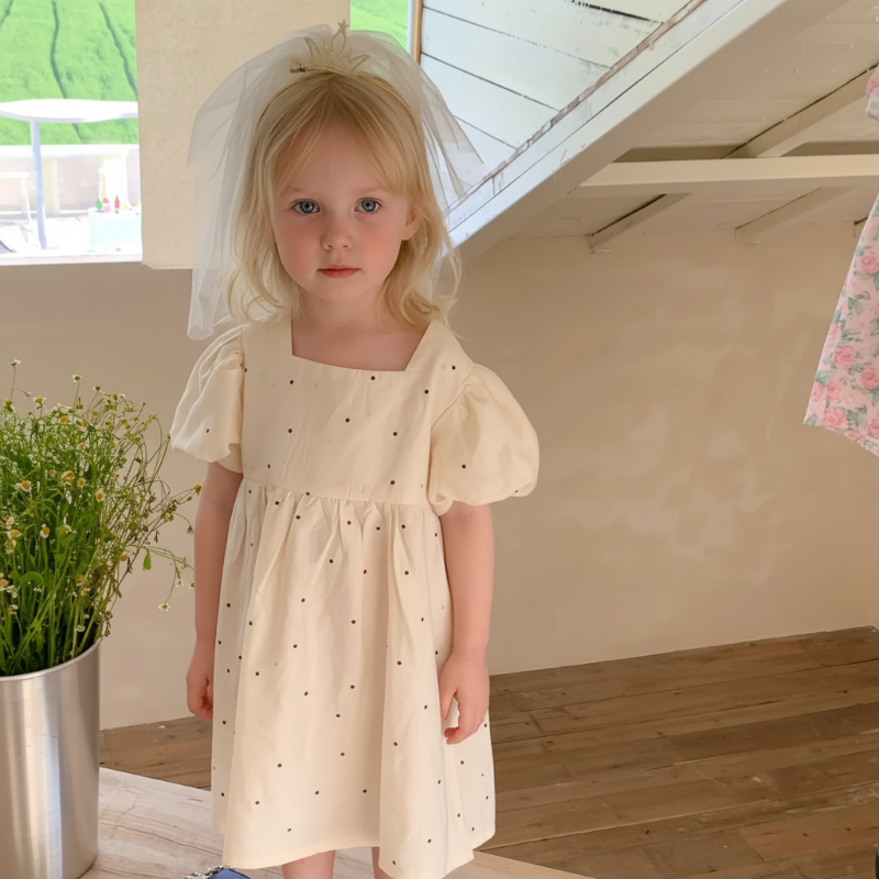 Front view of a child wearing the white dotted dress beside a flower-filled vase.