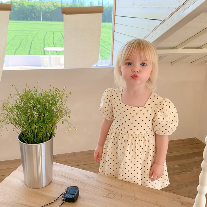 Child standing behind a wooden table in a white dotted dress with short puff sleeves.