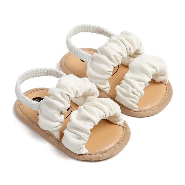 White open-toe baby sandals arranged at an angle, showing the ruched straps and tan footbeds