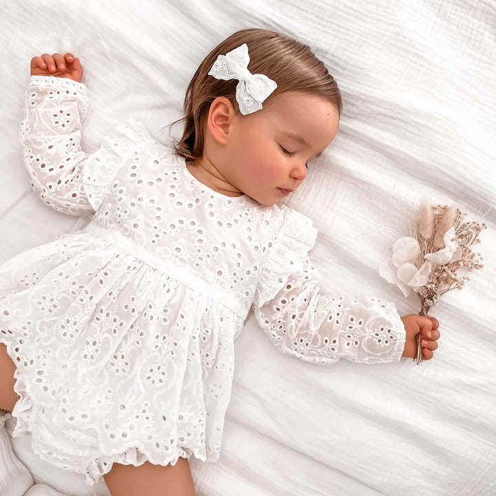Baby wearing a white eyelet ruffle top and matching shorts while lying on a bed.