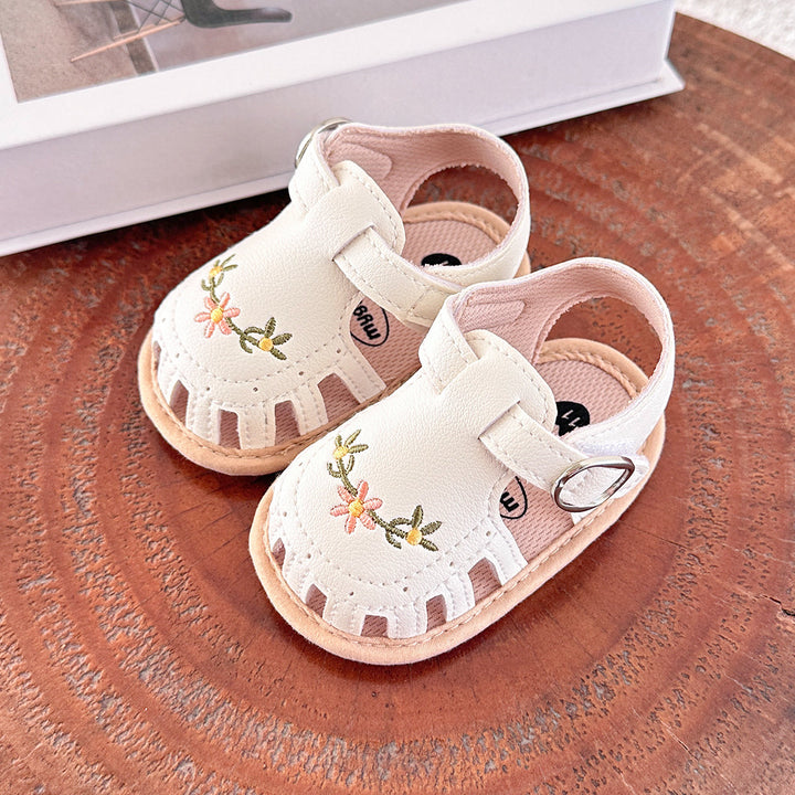 White baby sandals with floral embroidery and T-straps on a wooden table