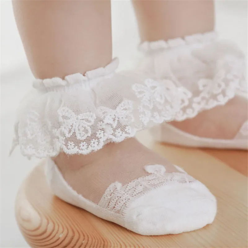 White baby socks with floral lace cuffs and sheer mesh uppers, worn on a wooden stool.
