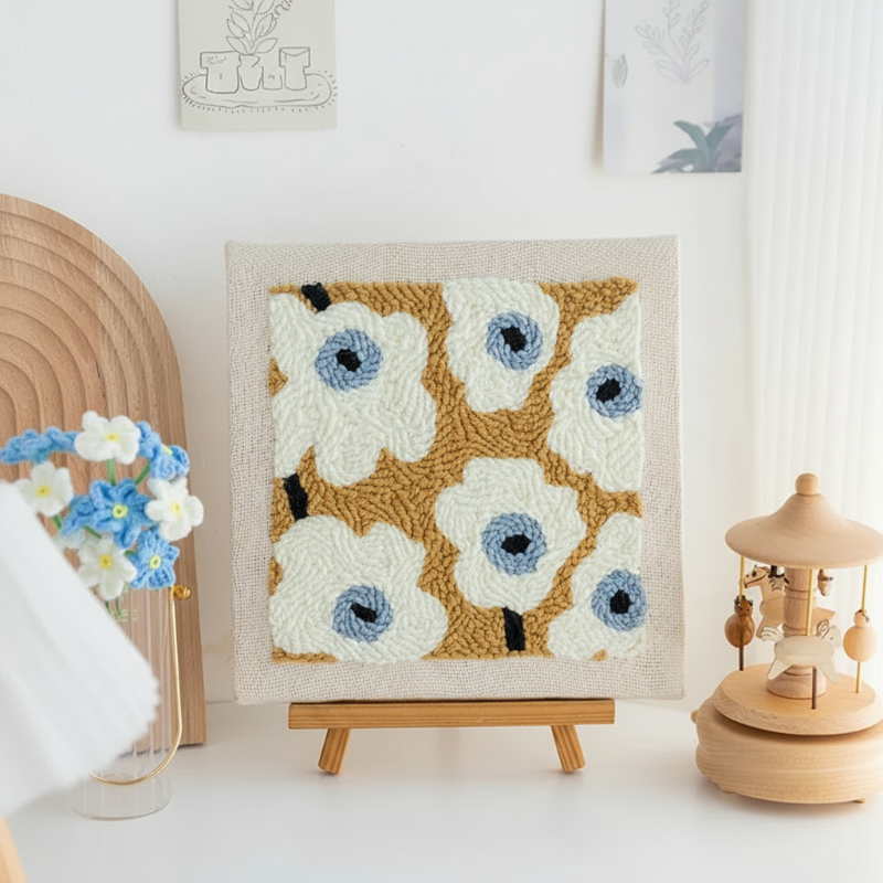Square white flower embroidery with blue centers displayed on a tabletop easel.