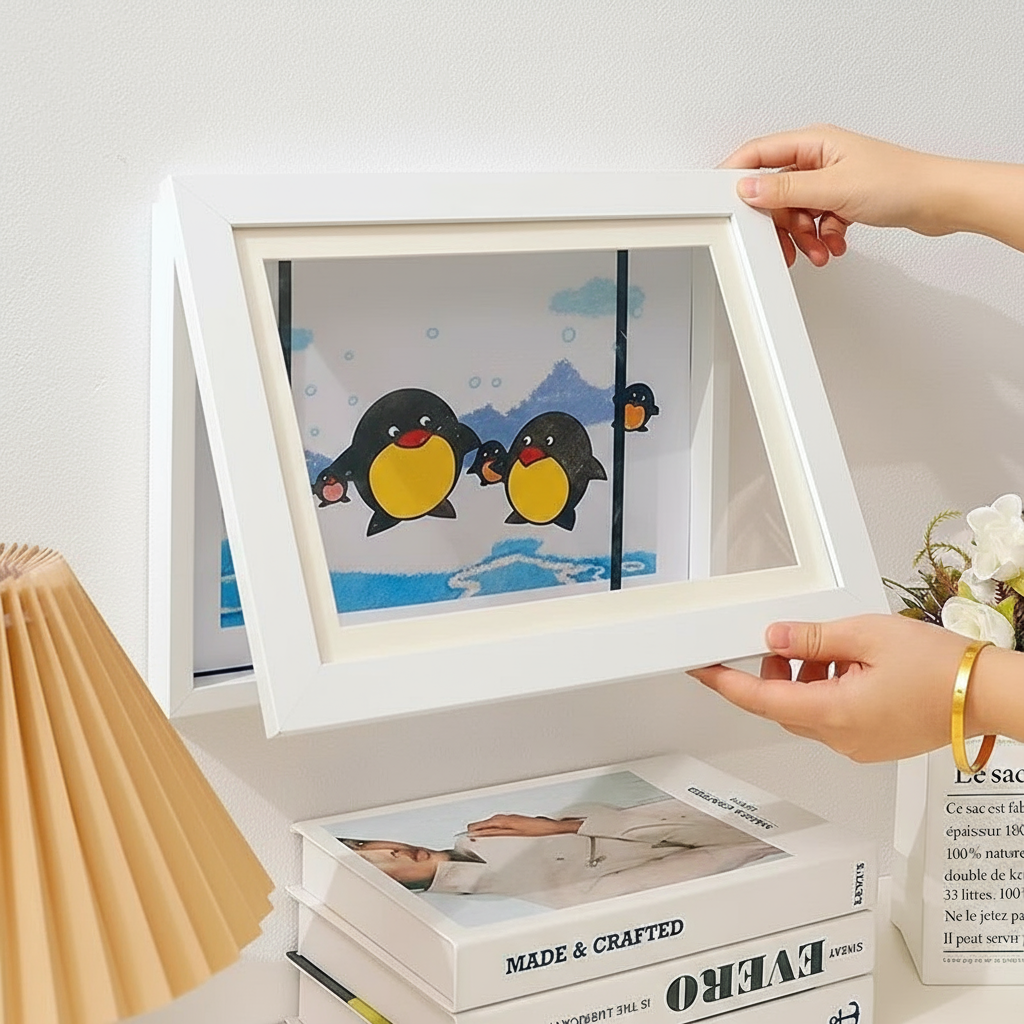 Hands opening a white art frame displaying a penguin drawing