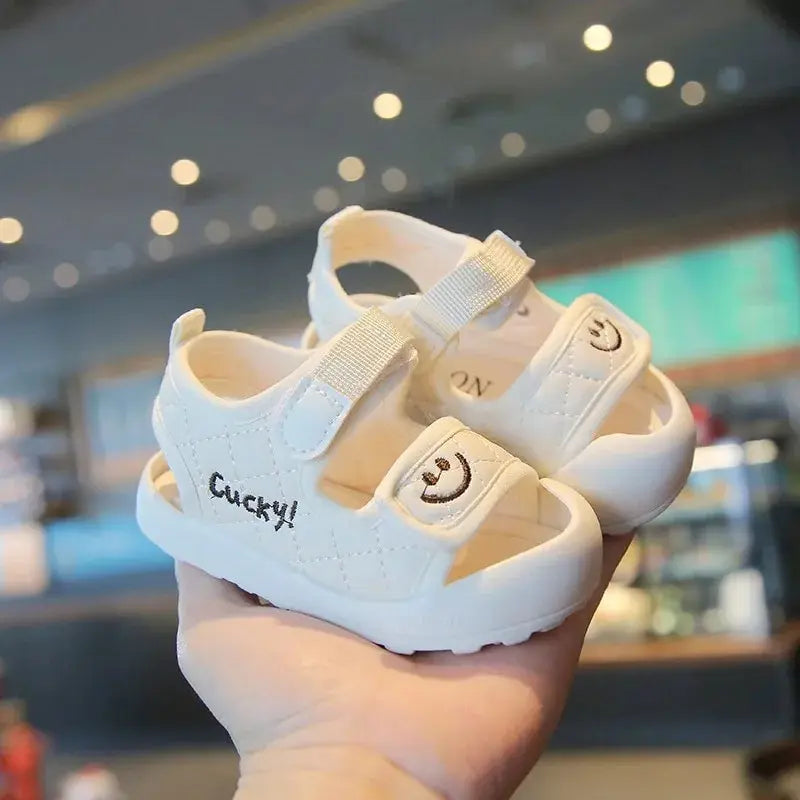 White children's sandals held in one hand, with smiley-face straps and rounded soles.