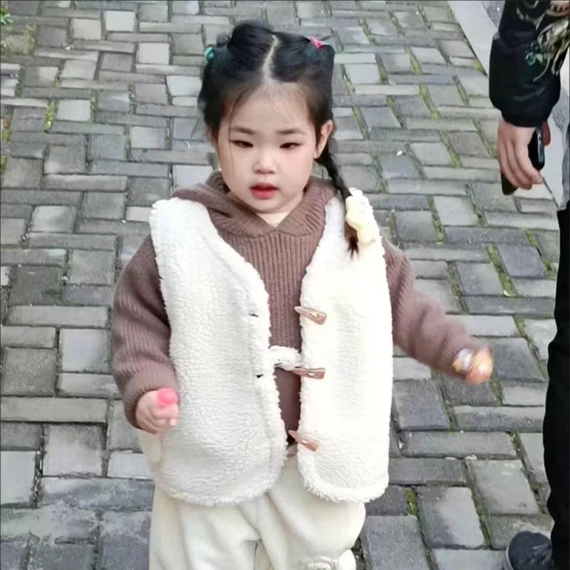 Child wearing a white textured vest over a brown sweater with cream pants outdoors