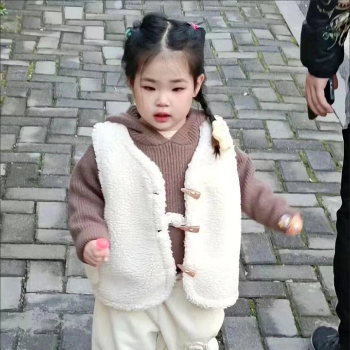 Child wearing a white textured vest over a brown sweater with cream pants outdoors
