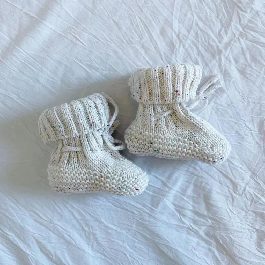 White knitted baby booties with folded ribbed cuffs and ankle ties on white fabric.