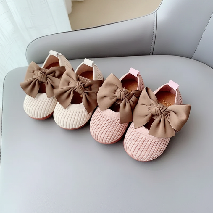 White and pink knitted baby shoes with oversized brown bows on a gray chair