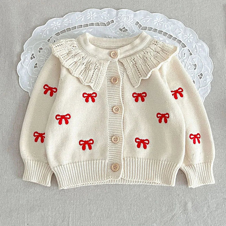 White button-front knit cardigan with red bow motifs, a ruffled collar, and ribbed cuffs