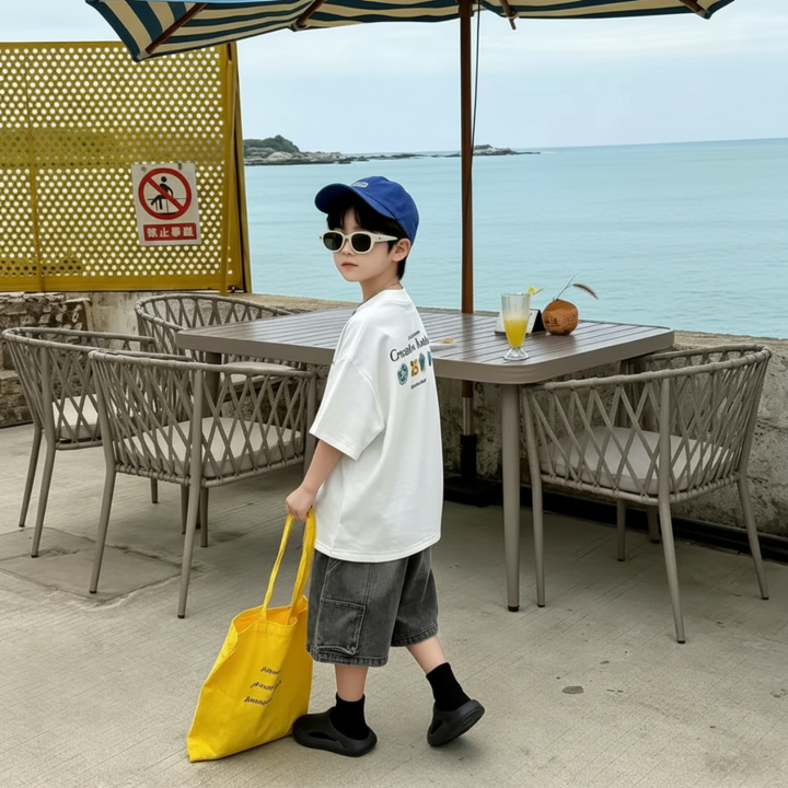 Boy in a white graphic T-shirt carrying a yellow tote beside outdoor cafe seating