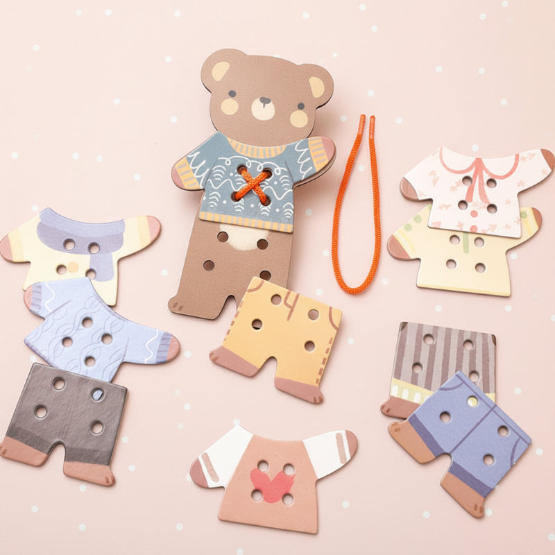 Wooden bear board, colorful clothing pieces, and an orange threading cord arranged on a pink background.