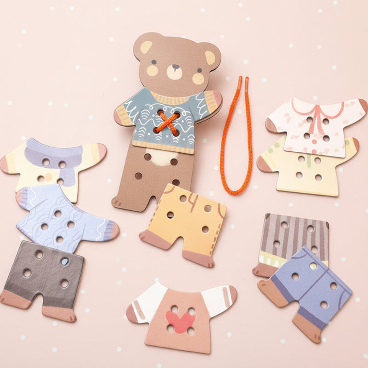 Wooden bear board, colorful clothing pieces, and an orange threading cord arranged on a pink background.