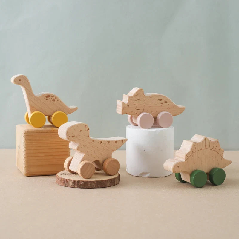 Four wooden dinosaur toy cars with yellow, green, pink, and natural wood wheels displayed on blocks