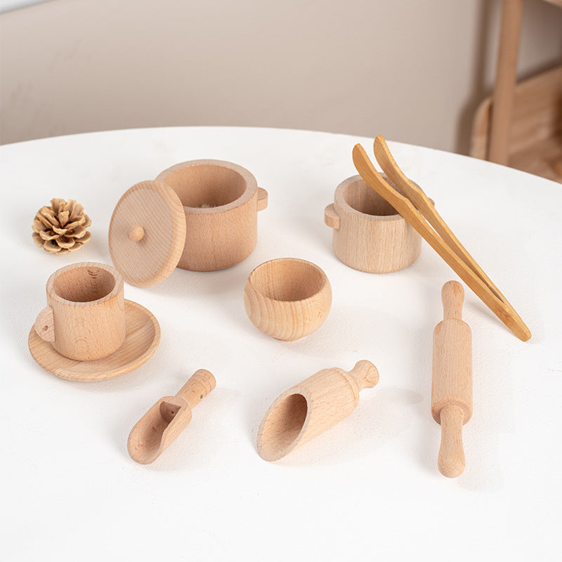 Wooden kitchen play set with cups, a bowl, plate, scoops, tongs and rolling pin on a white table.