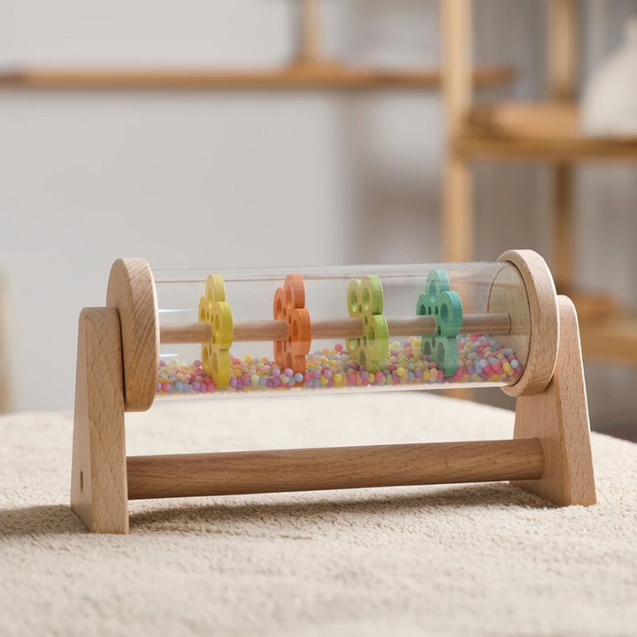 Wooden rain sound toy with a clear bead-filled cylinder resting on a cream rug.