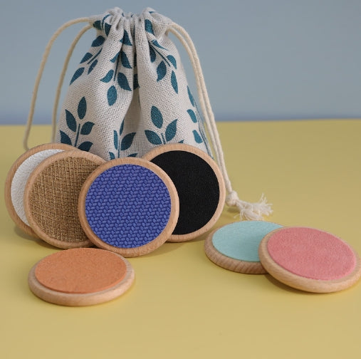 Seven wooden tactile discs with colored surfaces beside a leaf-print drawstring storage bag