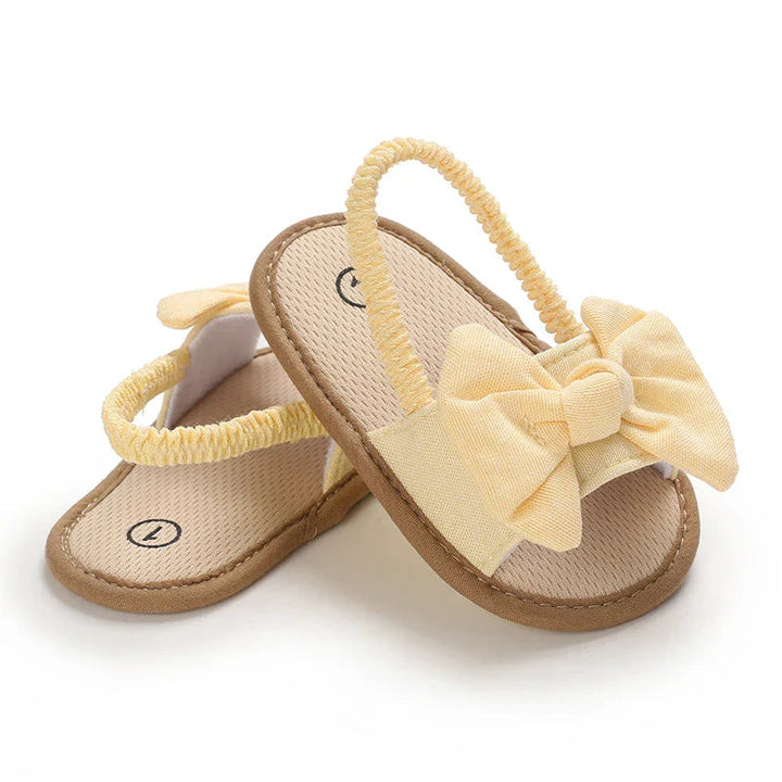 Yellow bow baby sandals arranged in a crossed pair with elastic heel straps